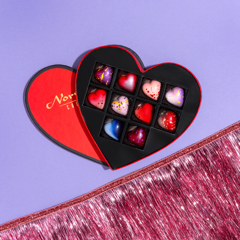 10 beautifully crafted heart-shaped chocolates in a red heart-shaped gift box. The chocolates are a variety of purple, pink and blue hearts. The box is offset on a purple background with a festive pink fringe ribbon in the bottom right corner. 